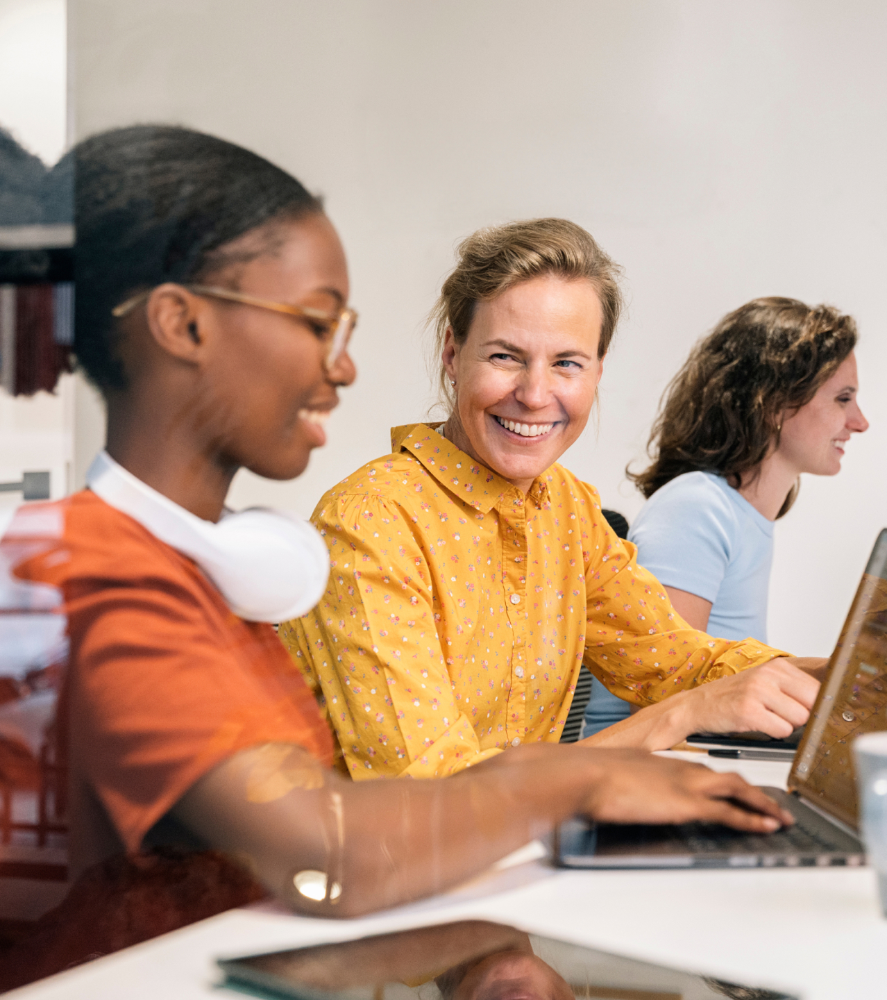 Drei Frauen arbeiten gemeinsam am Laptop im Büro, lächelnde Frau in gelber Bluse im Zentrum, diverse Teamarbeit in entspannter Atmosphäre