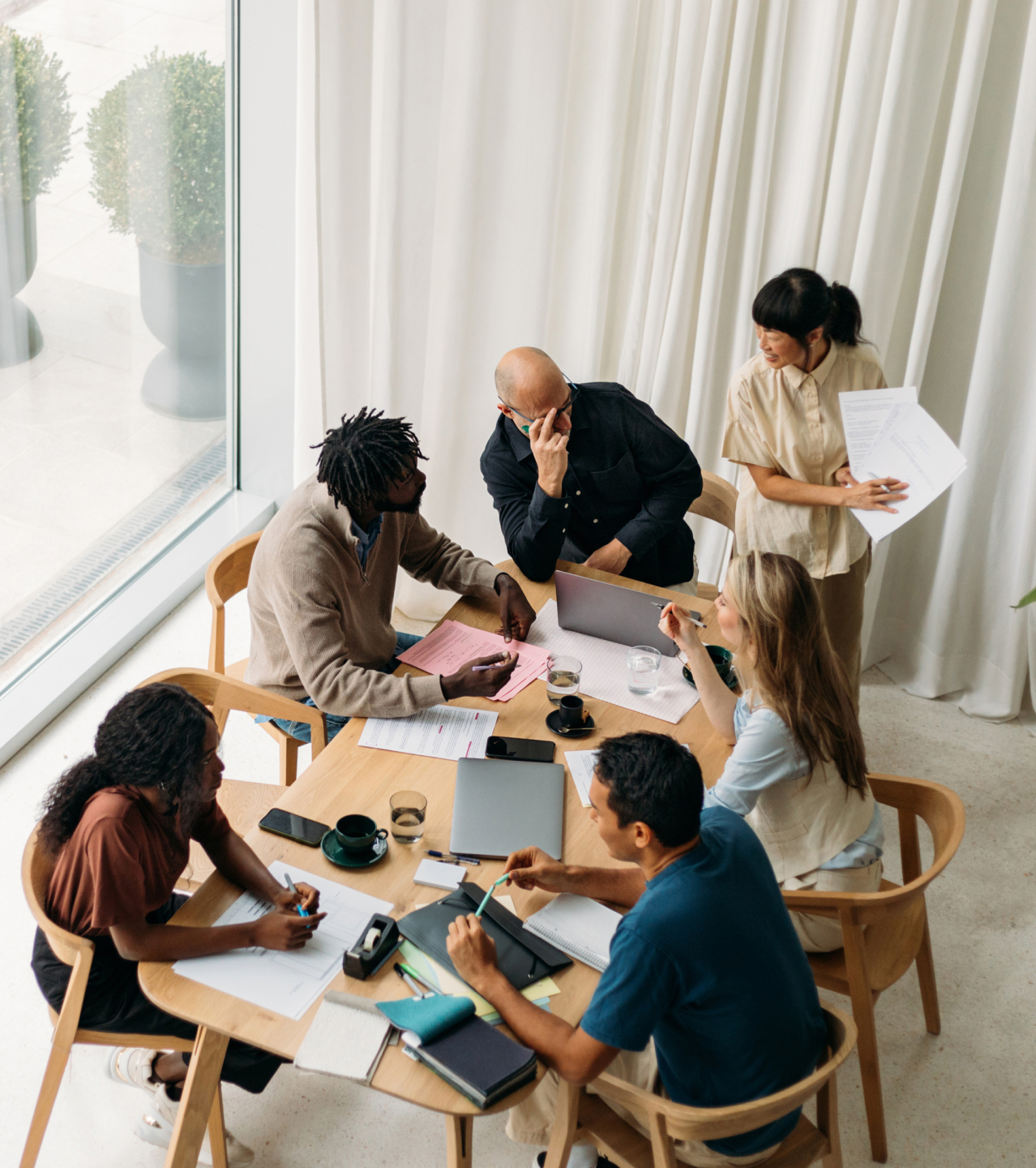 Sechsköpfiges diverses Team bei Besprechung an rundem Holztisch von oben fotografiert, mit Laptops, Dokumenten und Kaffeetassen in hellem minimalistischem Büro