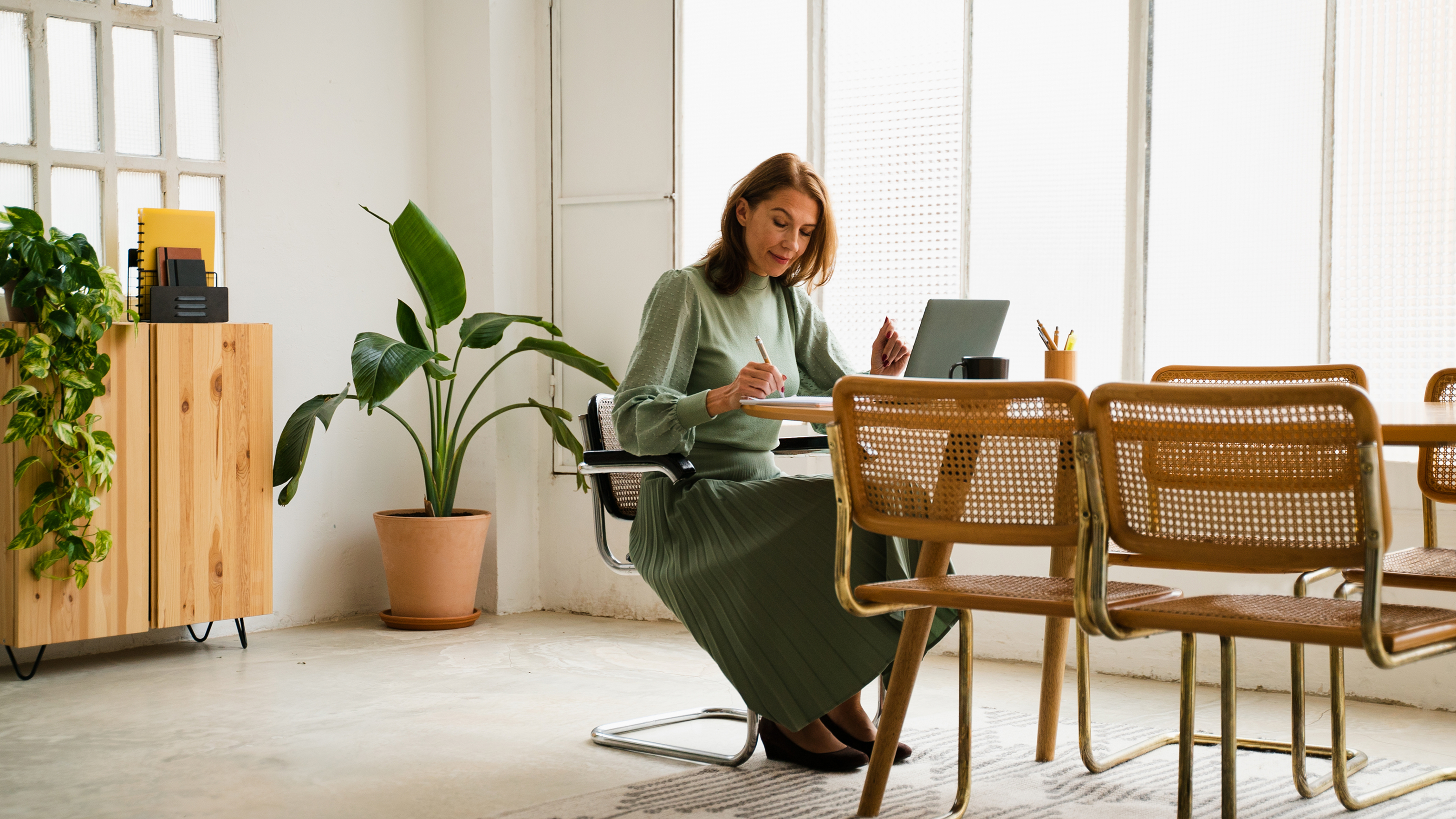 Frau in olivgrünem Outfit sitzt konzentriert am Schreibtisch und schreibt Notizen, Laptop und Stiftehalter vor sich, Rattan-Stühle im Vordergrund, helles Büro mit großen Fenstern, Grünpflanzen und Holzkommode