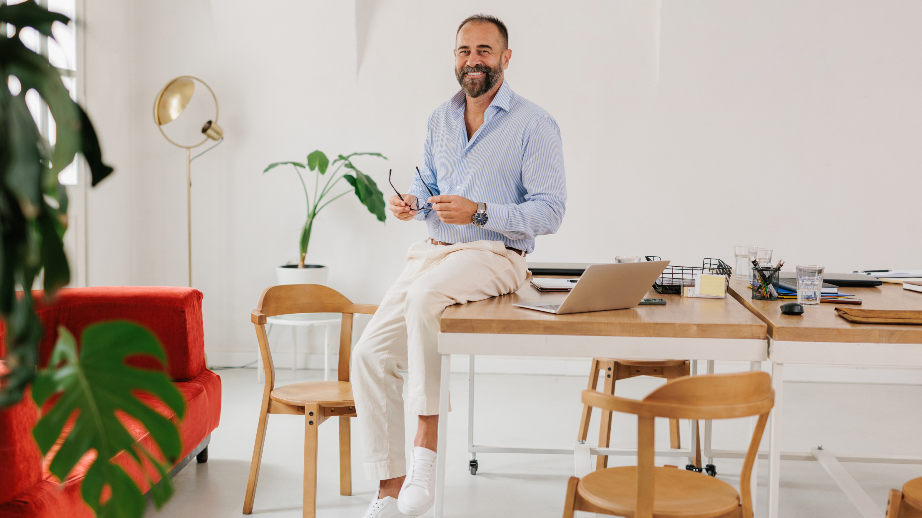 Lächelnder Geschäftsmann mit Bart in blau-weiß gestreiftem Hemd und weißer Hose sitzt entspannt auf Schreibtisch mit Brille in der Hand, helles minimalistisches Büro mit Holzmöbeln, goldener Stehlampe, rotem Sessel und Grünpflanzen