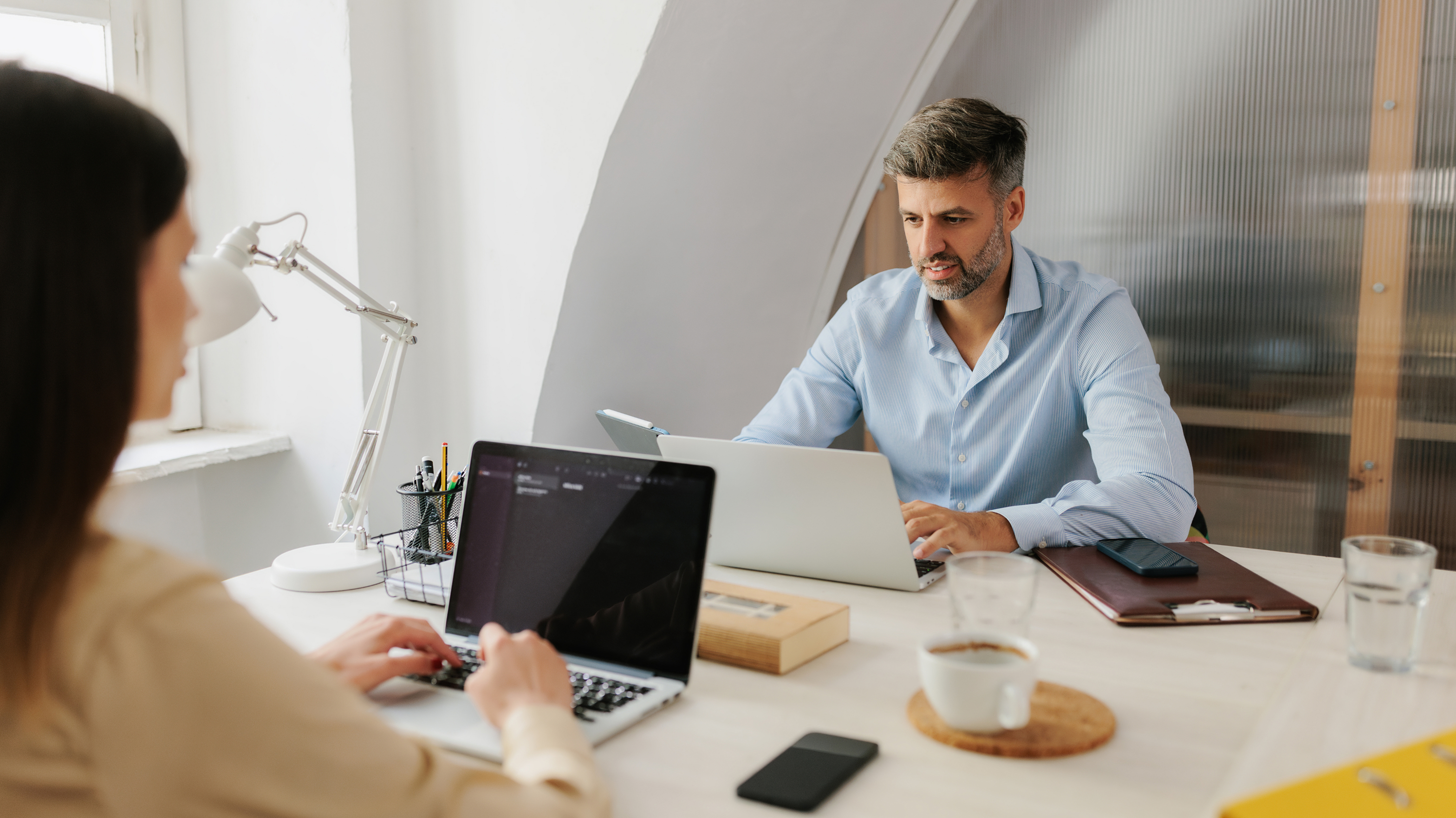 Zwei Kollegen arbeiten konzentriert mit Laptops an hellem Schreibtisch, Mann in hellblauem Hemd gegenüber von Frau in beiger Kleidung, Schreibtischlampe, Stifthalter, Kaffeetassen und Smartphone auf dem Tisch, modernes Büro mit Glaswand