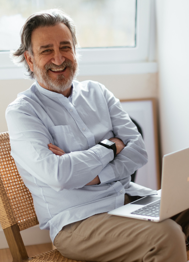 Lächelnder Geschäftsmann mittleren Alters mit grauem Bart in weißem Hemd sitzt entspannt mit verschränkten Armen vor Laptop in hellem Büro mit Grünpflanze und großem Fenster
