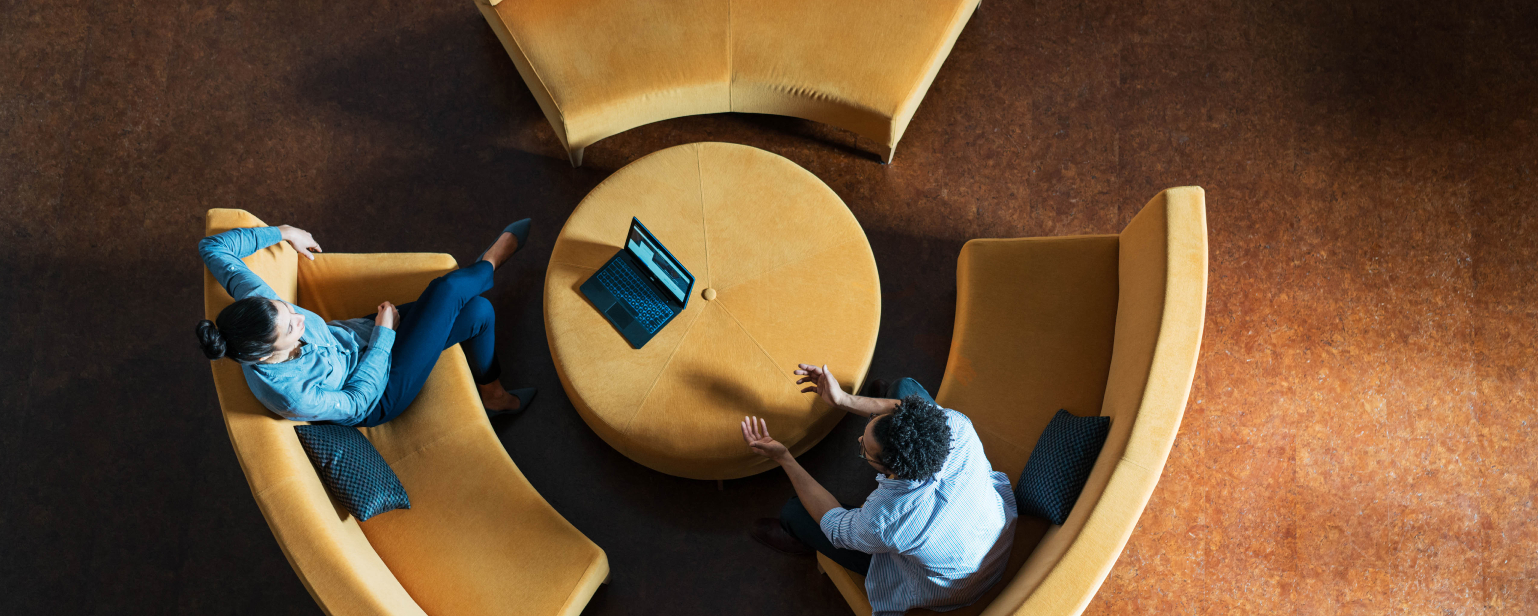 Vogelperspektive auf zwei Personen, die in einer modernen Lounge mit gelben Sofas an einem runden Tisch mit Laptop ein vertrauliches Beratungsgespräch führen.